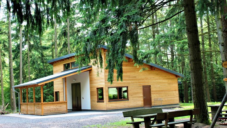 Ein modernes Holzhaus steht im Grünen, umgeben von dichten Bäumen. Vor dem Haus gibt es eine Terrasse und einen Sitzbereich.