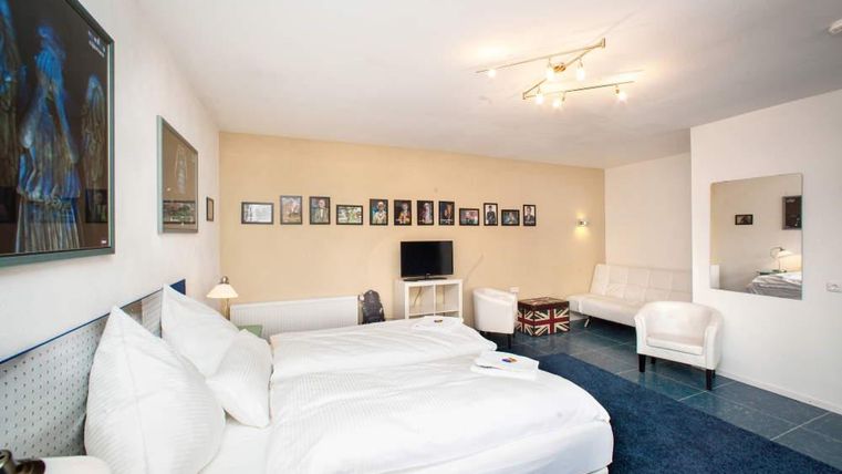 A modern hotel room with two single beds and elegant furnishings. Various pictures hang on the walls, and there is a television as well as a cozy seating area.