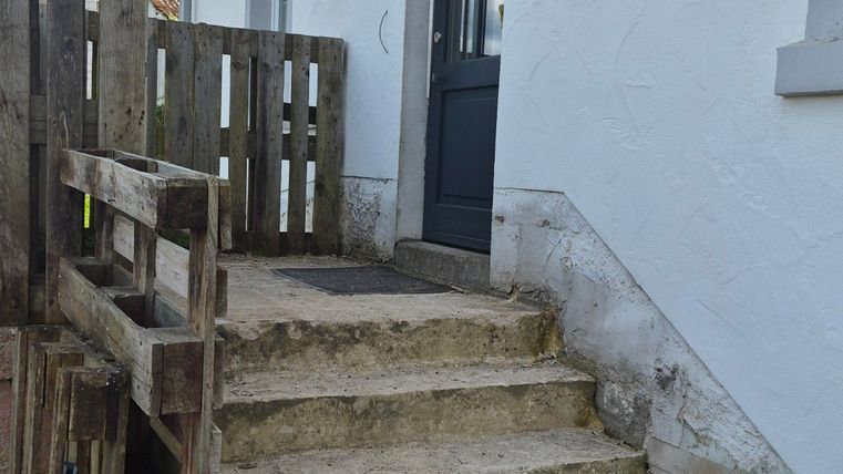 Een stenen trap leidt naar een donkere deur in een wit gebouw. Links is een houten hek zichtbaar, en de lucht is bedekt met enkele wolken.