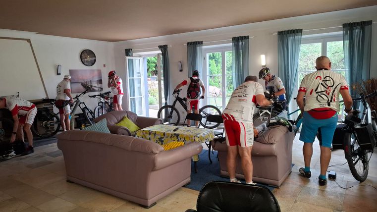 Eine Gruppe von Radfahrern bereitet sich in einem Wohnzimmer vor. Verschiedene Fahrräder sind im Raum verteilt und die Sportler tragen Rennkleidung.