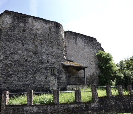 Ruine der Burg Bettingen mit massiven Steinmauern, umgeben von grüner Vegetation unter blauem Himmel., © TI Bitburger Land