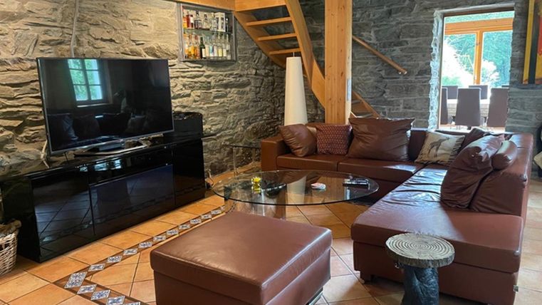 A cozy living space with a brown couch and a glass table. The room has stone walls and large windows that let in plenty of daylight.