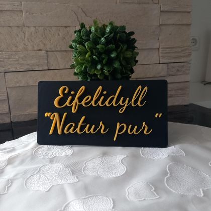 Een bord met de tekst "Eifelidyll - natuur puur" staat op een tafel. Op de achtergrond is een klein plantje te zien.