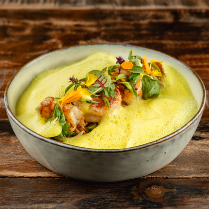 An elegant dish in a bowl, garnished with a green foam topping and colorful vegetables. The composition appears fresh and appealing.