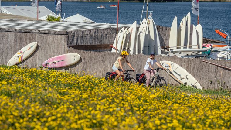 Deux personnes roulent à vélo le long d'un lac, entourées de fleurs jaunes et de planches de surf.