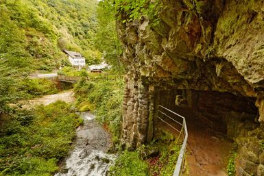 Eine natürliche Landschaft mit einem kleinen Bach und einer Felsenwand. Im Hintergrund sind Bäume und ein Gebäude sichtbar.