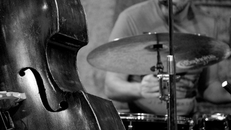 A double bass in the foreground and a drummer in the background. The scene conveys a relaxed musical atmosphere in a live setting.