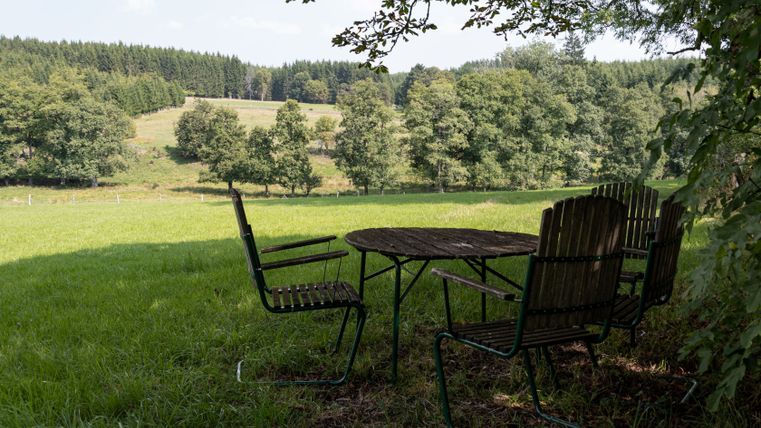 Une aire de repos avec une table en bois et des chaises sur une pelouse entourée d'arbres, avec vue sur un paysage boisé.