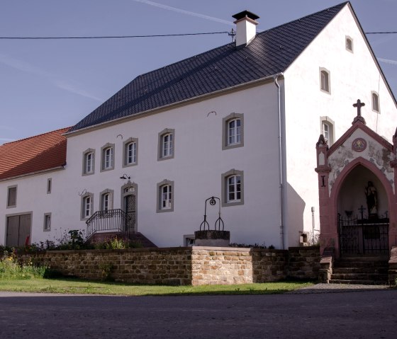 Une vieille ferme à la façade blanche et au toit rouge, à côté d'une petite chapelle avec une croix. Le ciel est bleu et clair., © Tourist-Information Bitburger Land
