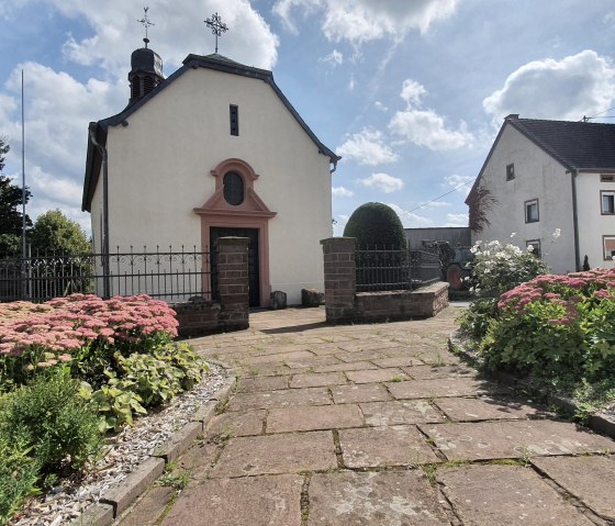 Église de village à Sefferweich, entourée de parterres fleuris et d'un chemin pavé, sous un ciel bleu avec des nuages., © Tourist-Information Bitburger Land