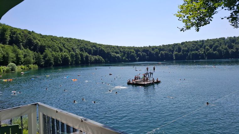 A picturesque lake surrounded by green forests. People are swimming and playing on a platform in the water.