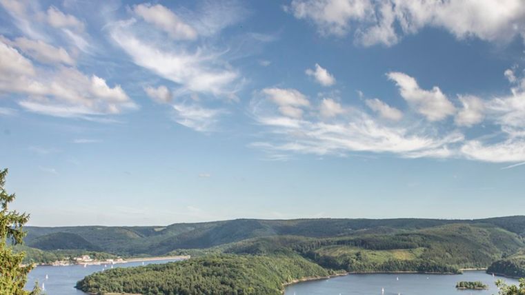 A picturesque lake surrounded by green hills and wooded shores. The sky is blue with some clouds.