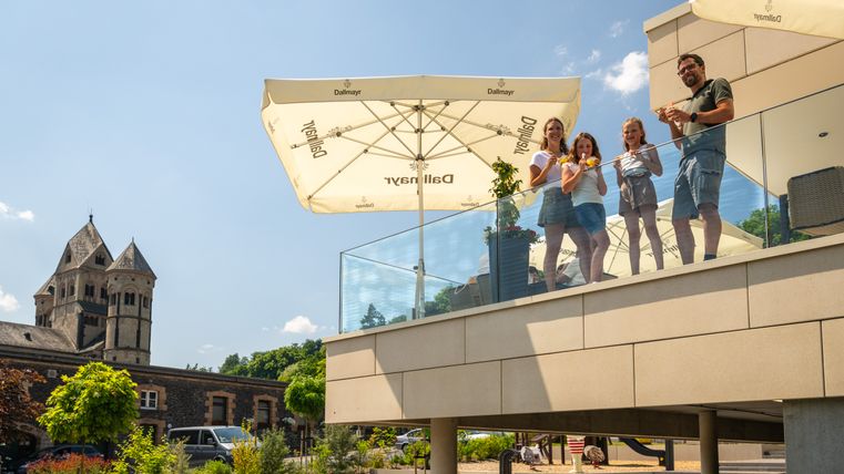 Eine vierköpfige Familie mit Eis in der Hand auf einer erhöhten Terrasse eines Cafés. Im Hintergrund das Kloster 