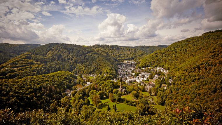 Panoramic view in cloudy weather of the town of Bad Bertrich, surrounded by a hilly, densely wooded landscape.