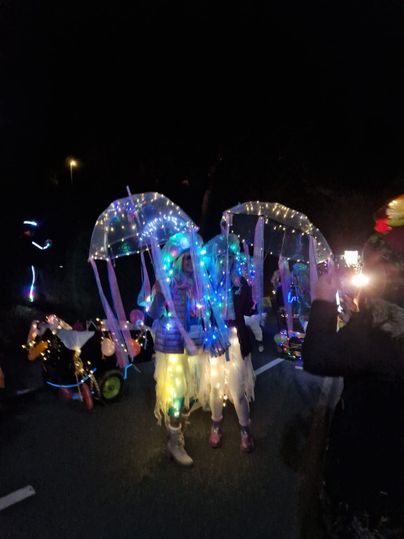 Eine bunte Parade mit beleuchteten Schirmen und festlich gekleideten Menschen. Die Atmosphäre ist lebhaft und fröhlich, trotz der Dunkelheit.