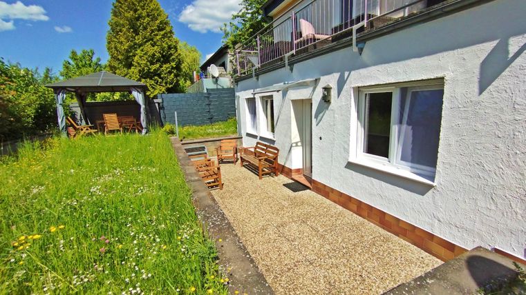 Un jardin ensoleillé avec une terrasse et des sièges en bois. À l'arrière-plan, on voit des arbres et un pavillon.