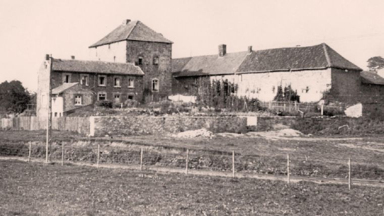 A historic farmhouse with a large, landscaped garden. Other buildings are visible in the background, surrounded by a rural setting.