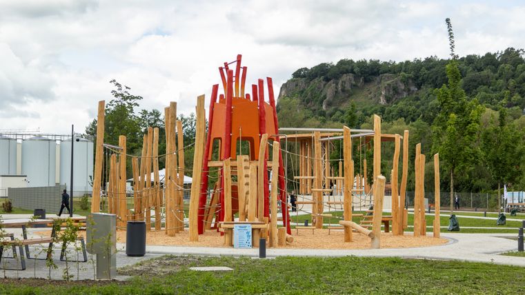 Een moderne speeltuin met een houten klimrek en rode elementen, omgeven door een groen landschap en heuvels op de achtergrond.
