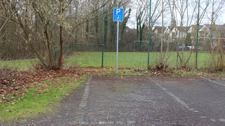 Ein Parkplatz mit einem behindertengerechten Parkplatzschild, dahinter ein eingezäunter Fußballplatz