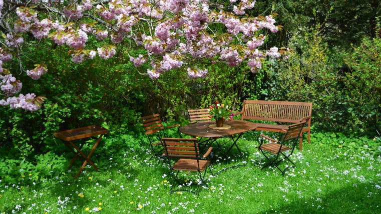 Ein schöner Garten mit einem Holztisch und Stühlen, umgeben von bunten Blumen. Über dem Tisch blüht ein Baum mit rosa Blumen.