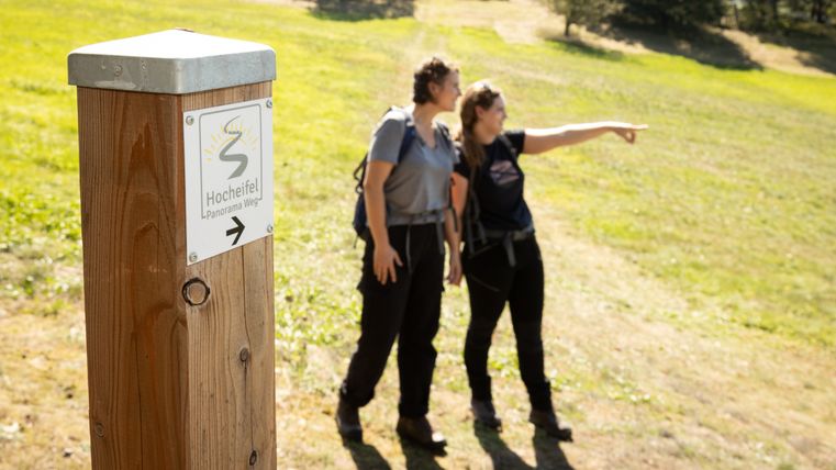 Twee mensen op een wandelpad in de Hocheifel, één wijst in de verte. Een wegwijzer met het opschrift 'Hocheifel Panorama Weg'.