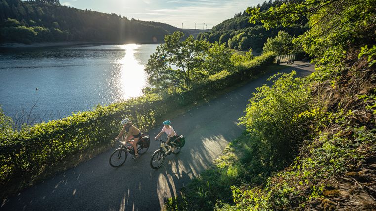 Twee fietsers rijden op een pad langs een meer in de zon, omringd door groene natuur.