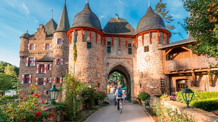 Kasteel Satzvey met fietsers op de voorgrond, omringd door bloeiende rozen en een blauwe lucht.