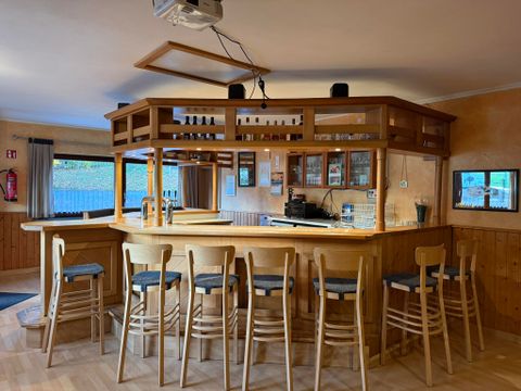 Een gezellige bar met houten wanden en hoge stoelen. Achter de bar zijn planken met dranken en een koffieautomaat zichtbaar.