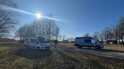 Deux camping-cars se trouvent sur une place avec des arbres en arrière-plan. Le ciel est clair et ensoleillé.