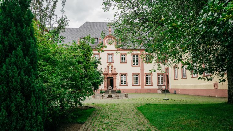 An impressive building with red and beige colors surrounded by trees and plants. The paved path leads to the entrance door of the house.