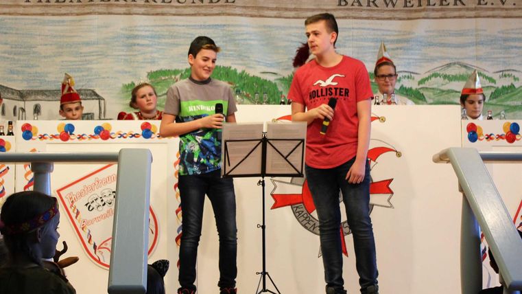 Two boys are standing on stage and singing with microphones. In the background, spectators are sitting in costumes.
