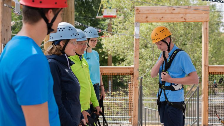 Un groupe de personnes se tient sur une plateforme et se prépare pour une activité d'aventure. Tout le monde porte des casques et des harnais de sécurité.