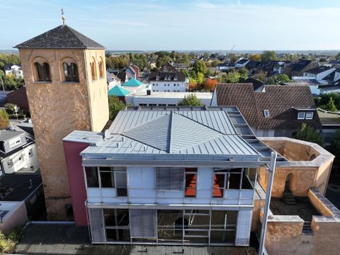 Ein modernes Gebäude mit einem alten Kirchturm im Hintergrund. Die Umgebung besteht aus Wohnhäusern und Bäumen.
