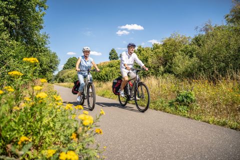 Two cyclists are riding on a bike path through a green landscape. The sun is shining, and the sky is blue with a few clouds.