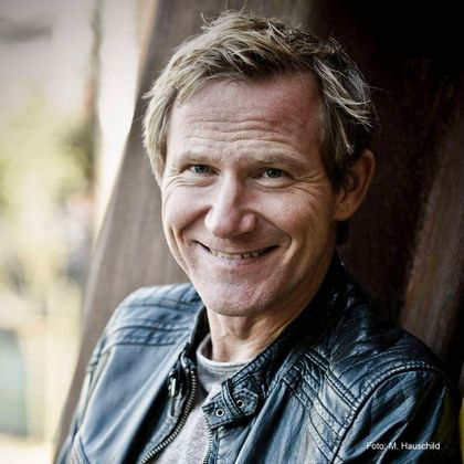 A smiling man in a leather jacket in front of a blurred background landscape. He has short, blonde hair and a friendly face.