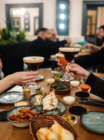 Deux verres de cocktails s'entrechoquent, tandis qu'une table dressée avec différents plats est visible. À l'arrière-plan, des invités joyeux se distinguent, profitant de l'atmosphère.