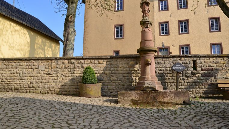 Een historische straat met een stenen fontein en een oude muur. Op de achtergrond zijn meerdere ramen van een gebouw zichtbaar.