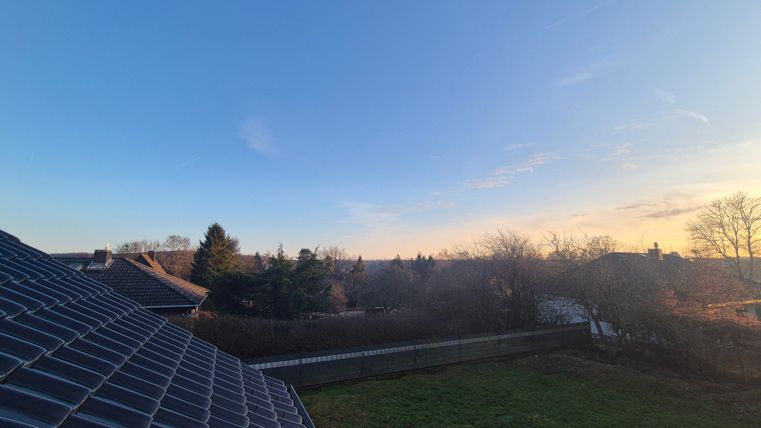 Une vue depuis un toit avec un ciel clair et des couleurs douces pendant le coucher de soleil. Au premier plan, il y a des arbres et un jardin.