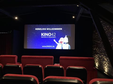 Een bioscoopzaal met rode stoelen en een groot scherm. Op het scherm staat "WELKOM BIJ KINO42".