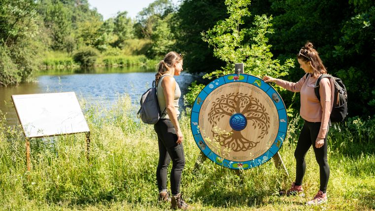 Twee vrouwen staan bij een decoratief houten wiel aan de oever van een meer.