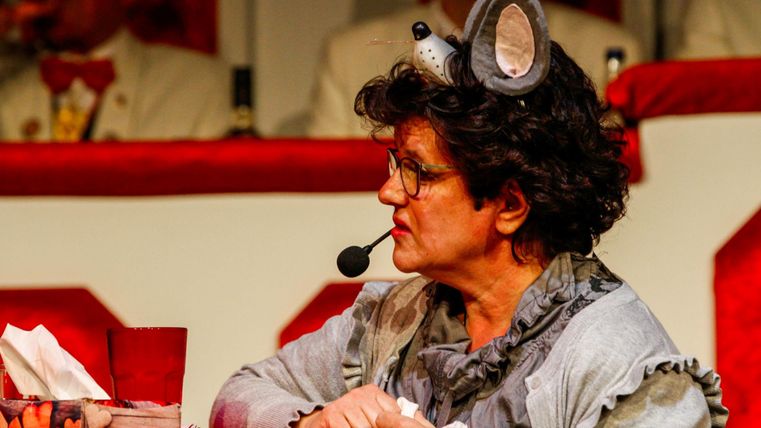 A woman with a mouse ears headband is speaking into the microphone. In the background, people in white suits can be seen.