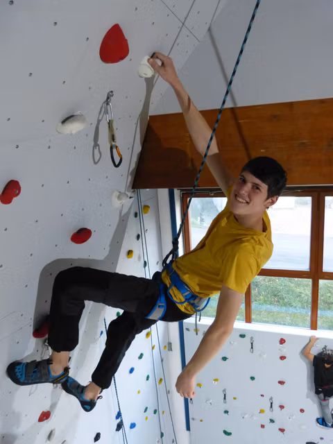Ein junger Kletterer hängt an einer Kletterwand und lächelt. Die Wand ist mit verschiedenen Griffen in unterschiedlichen Farben ausgestattet.