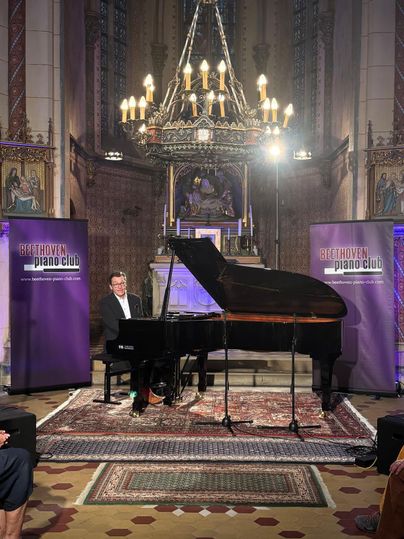 Ein Pianist spielt Klavier in einem festlichen Raum mit Kirchenarchitektur. Im Hintergrund sind Banner des Beethoven Piano Clubs zu sehen.