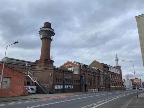 Ein urbaner Straßenzug mit historischen Gebäuden und einem markanten Aussichtsturm. Der Himmel ist bewölkt und die Straßen sind ruhig.