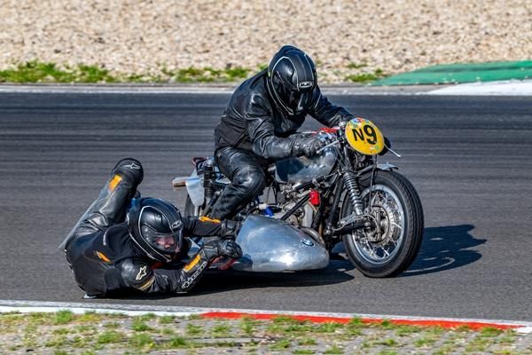 Een motorcoureur in lederen racepak en een passagier op een racefiets. De coureur bevindt zich in een bocht en toont een actievolle rijtechniek.