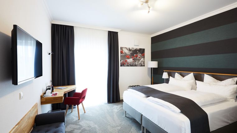 Ein modernes Hotelzimmer mit einem großen Bett und einem Arbeitsplatz. Helle Farben und große Fenster sorgen für eine angenehme Atmosphäre.