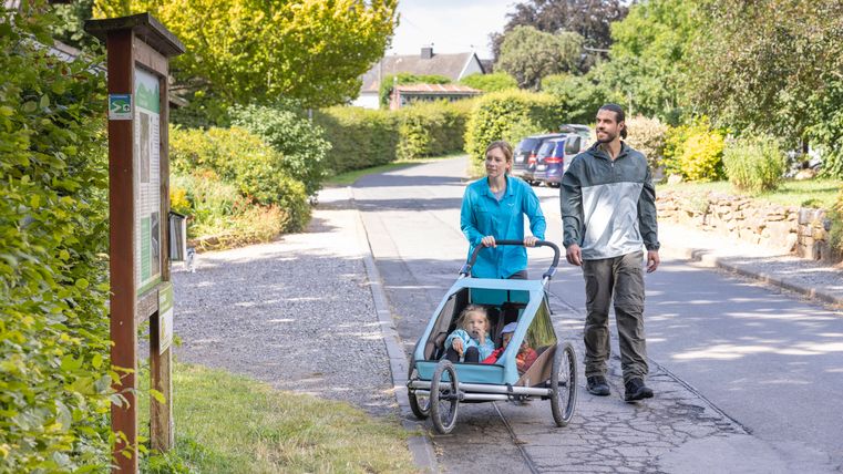 Een stel duwt een kinderwagen over een trottoir in een groene woonwijk.