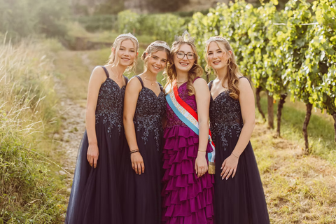 Four young women in elegant evening gowns pose smiling in a vineyard. The atmosphere is festive and cheerful.