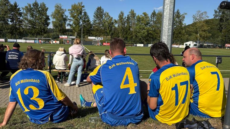 A group of soccer players is sitting on the ground and looking at the field. The players are wearing blue and yellow jerseys of DJK Mittenbach.