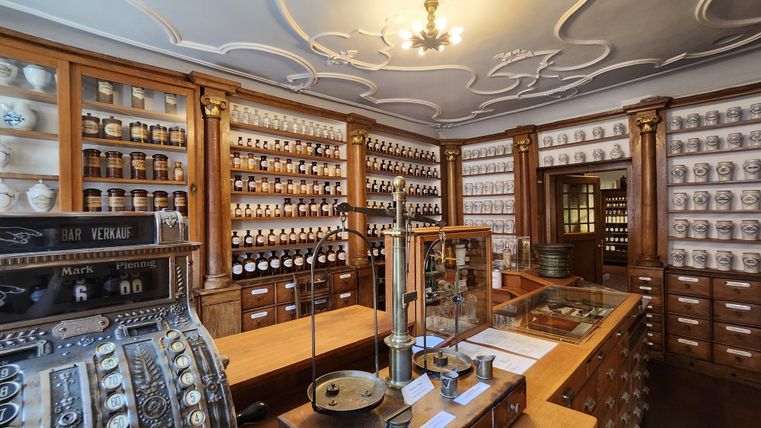 A traditional sales room with wooden shelves and many jars. In the foreground, there is an old scale and a cash register.